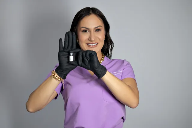Bianca in lilac scrubs and black gloves, holding a vial in front of her